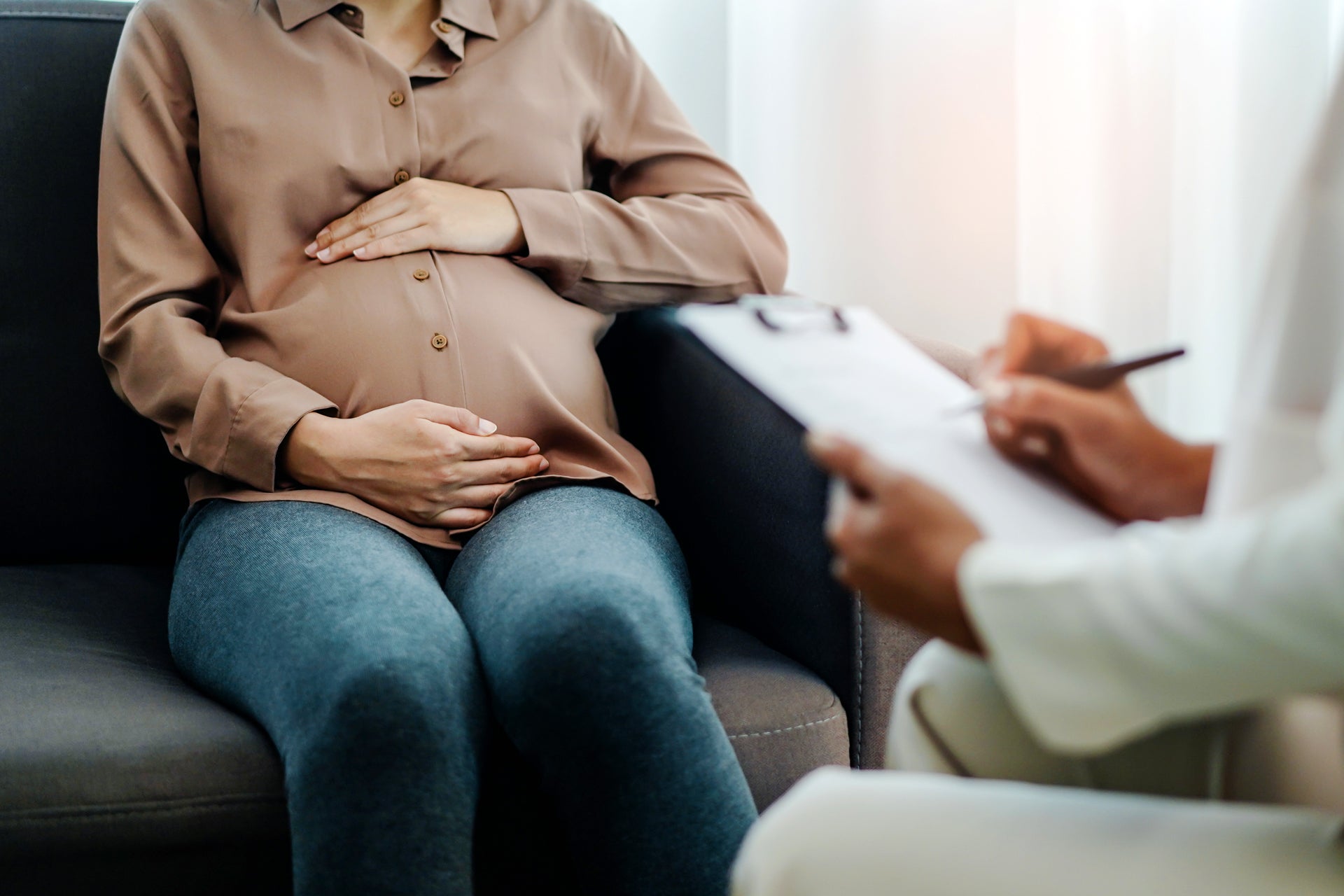 Eine schwangere Frau sitzt auf einem Sofa und hält ihre Hände auf den Bauch. Gegenüber sitzt eine weitere Person, die Notizen auf einem Klemmbrett macht, vermutlich im Rahmen eines Beratungsgesprächs oder einer Untersuchung.