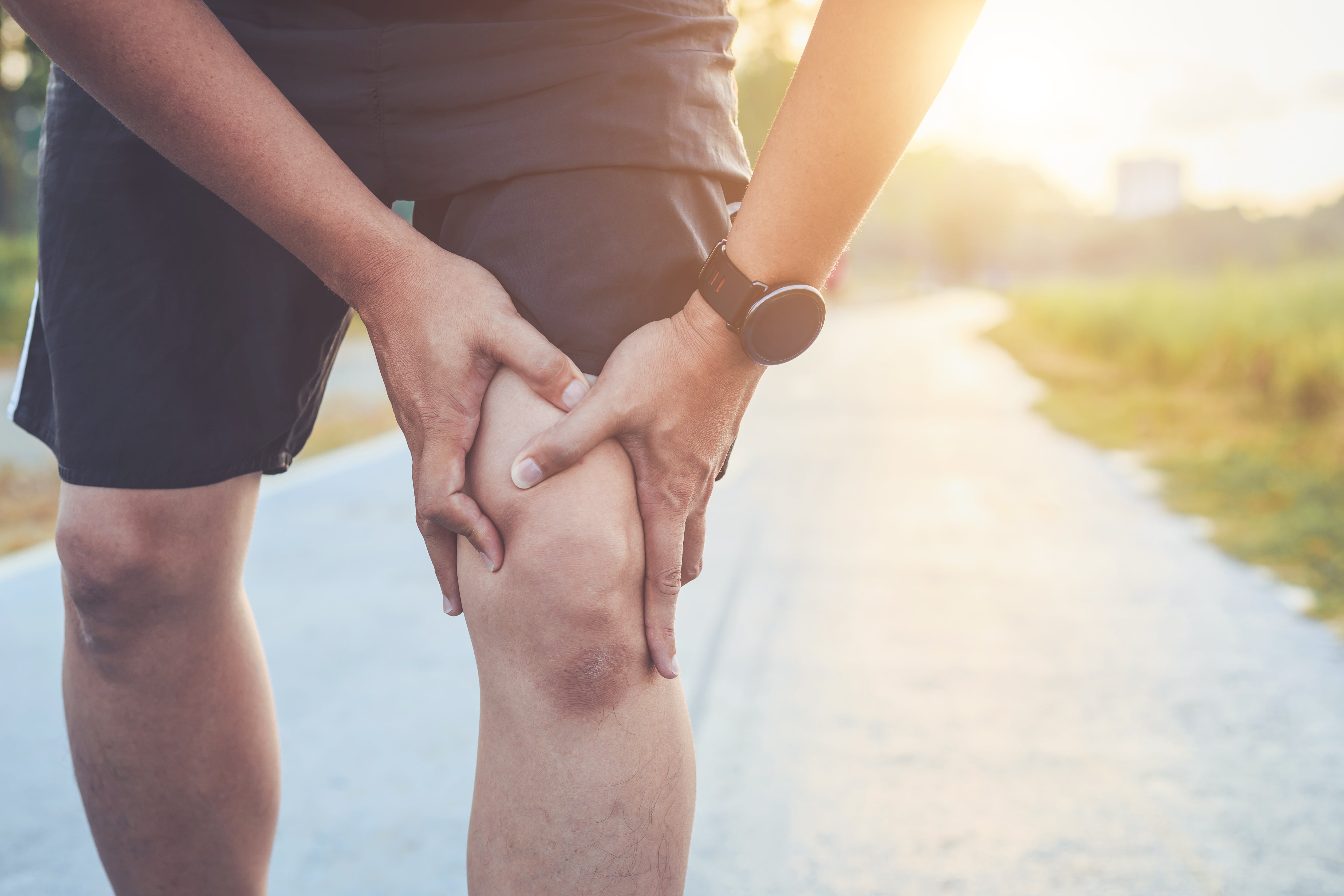 Abgebildet ist eine Person im Freien, die sich mit beiden Händen schmerzend an das Knie fasst. Die Körperhaltung und Mimik deuten auf Beschwerden oder Schmerzen im Kniebereich hin.