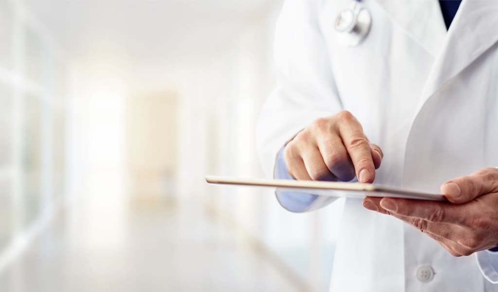 Abgebildet ist eine Person im Arztkittel, die in einer hellen Umgebung ein Tablet bedient. Der Fokus liegt auf den Händen und dem Gerät, der Hintergrund ist unscharf gehalten.