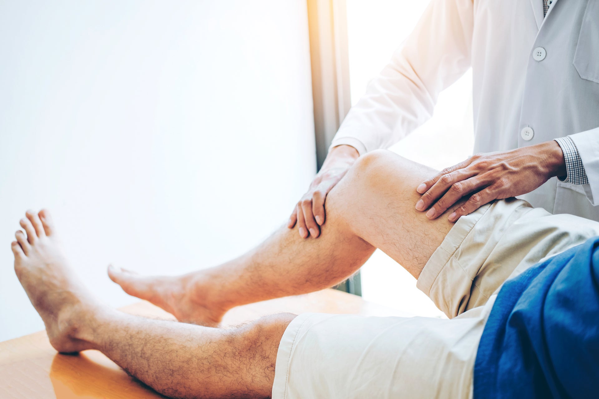 Auf dem Bild ist eine Situation in einer Arztpraxis oder physiotherapeutischen Behandlung zu sehen. Eine Person liegt auf einer Untersuchungsliege, während eine andere Person (vermutlich ein Arzt oder Physiotherapeut im weißen Kittel) das Bein bzw. Knie des Patienten mit beiden Händen untersucht oder abtastet. Der Fokus liegt auf dem Knie, was darauf hindeutet, dass dieses auf mögliche Verletzungen oder Beschwerden hin untersucht wird.