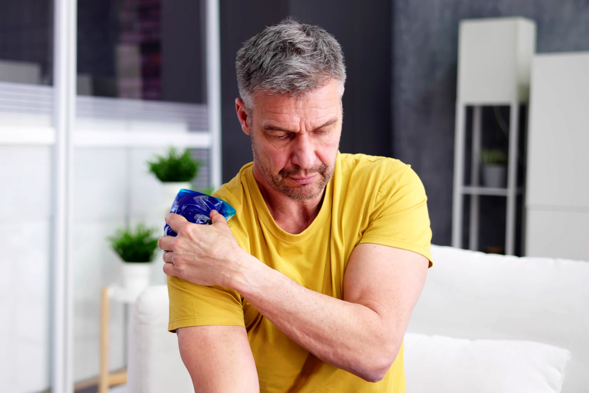 Auf dem Bild ist ein Mann zu sehen, der sich ein Kühlpack auf seine rechte Schulter hält. Er sitzt auf einem Sofa und trägt ein gelbes T-Shirt. Der Gesichtsausdruck wirkt konzentriert oder leicht schmerzhaft, was darauf hindeutet, dass er vermutlich Schmerzen oder eine Verletzung an der Schulter hat und diese mit Kälte behandelt. Der Hintergrund zeigt einen modernen Innenraum.