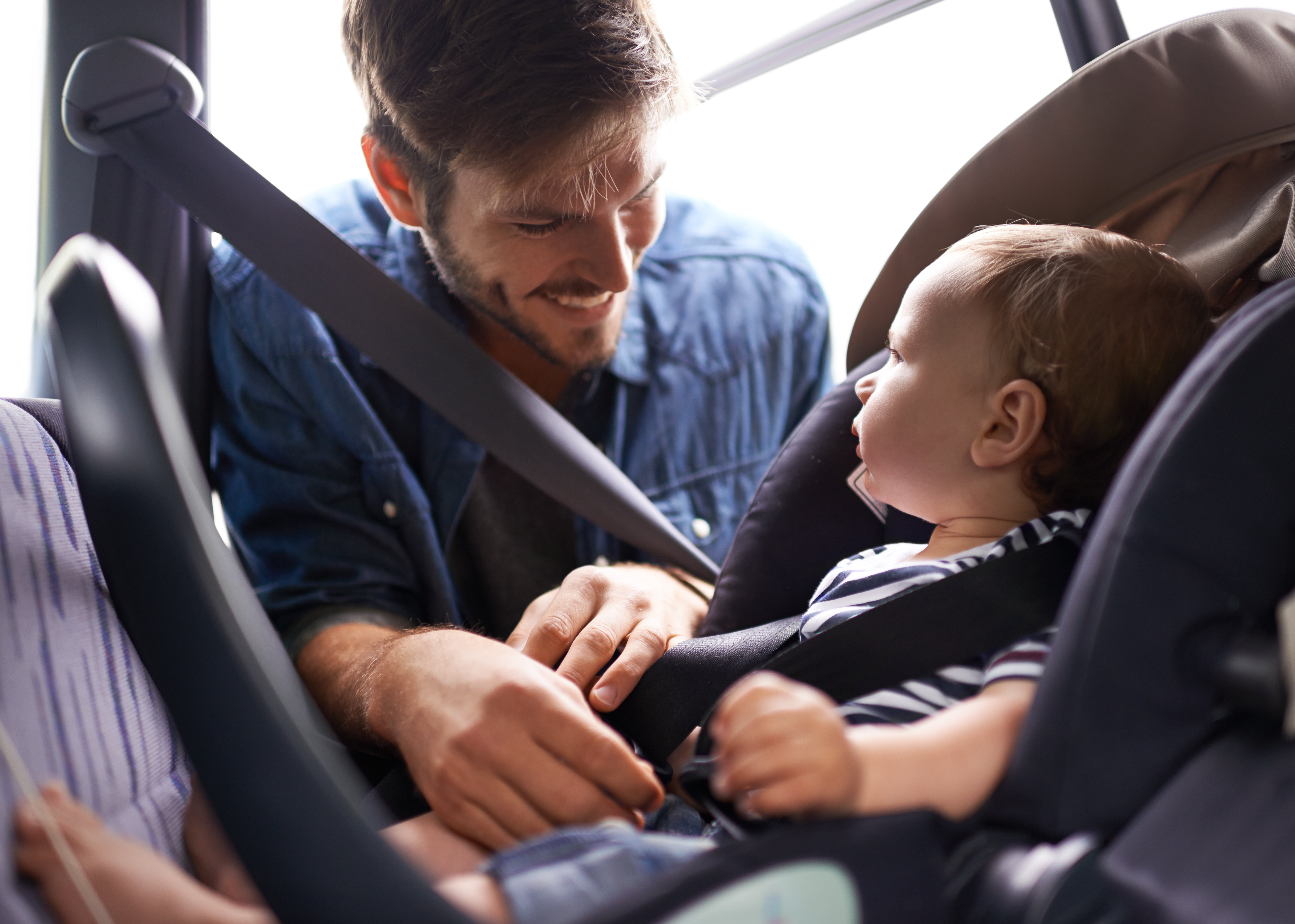 Auf dem Bild ist ein Mann zu sehen, der mit einem Baby in einem Auto sitzt. Der Mann lächelt das Baby an und hilft dabei, es im Kindersitz mit dem Sicherheitsgurt anzuschnallen. Die Szene wirkt liebevoll und fürsorglich und vermittelt den Eindruck von Sicherheit und Geborgenheit auf einer Autofahrt.