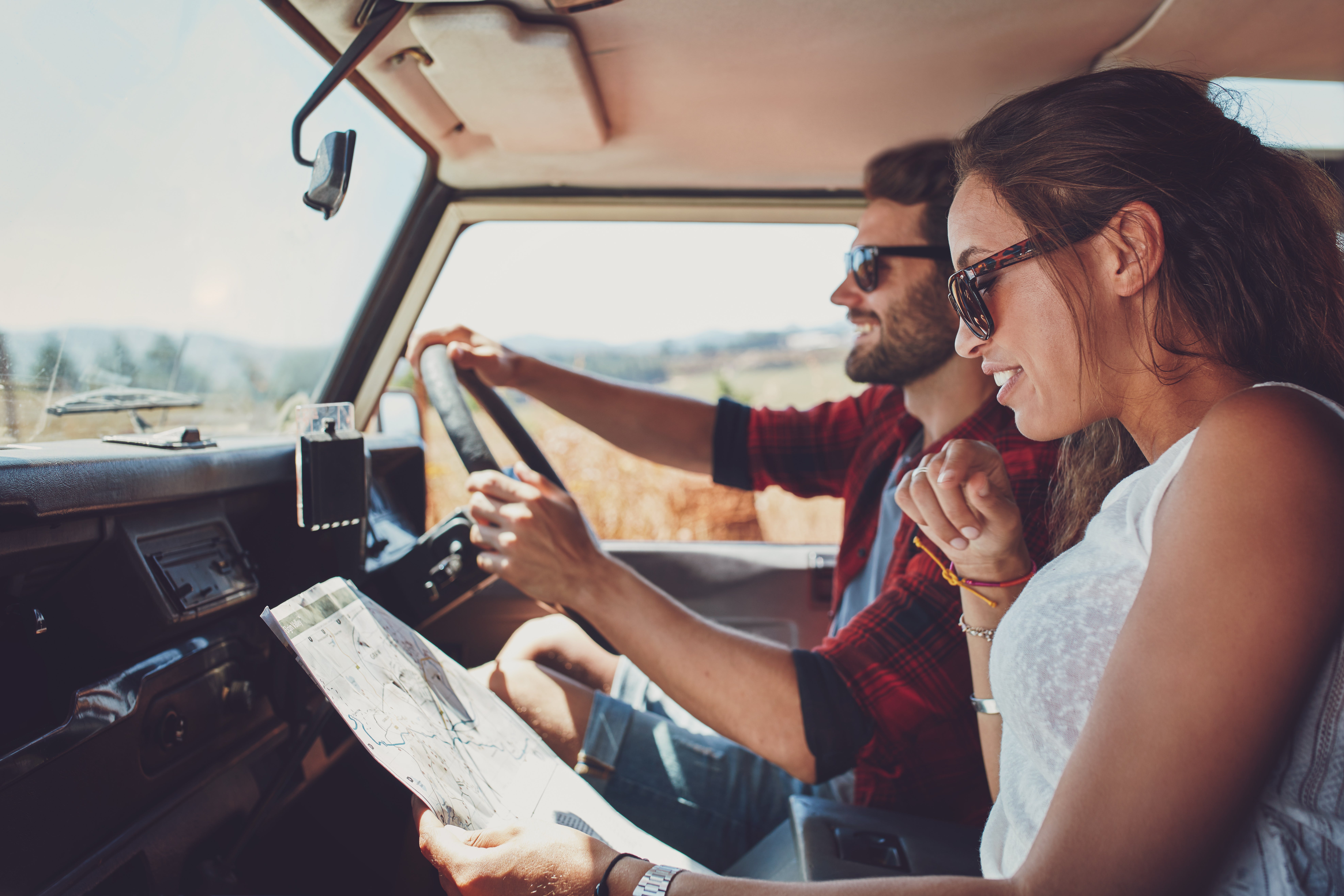Auf dem Bild sitzt ein junges Paar in einem Auto, vermutlich auf einer Reise oder im Urlaub. Der Mann fährt, während die Frau auf dem Beifahrersitz sitzt und eine Landkarte hält, in die sie konzentriert und zugleich fröhlich blickt. Beide tragen Sonnenbrillen und sind gut gelaunt. Die Umgebung draußen sieht ländlich und sonnig aus. Die Szene vermittelt Reiselust, Abenteuer und gemeinsame Planung unterwegs.