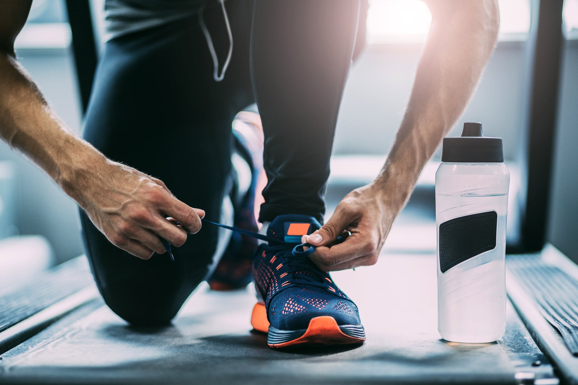 Auf dem Bild ist eine Person zu sehen, die auf einem Laufband steht und gerade ihre Sportschuhe zubindet. Fokus liegt auf den Händen, dem Schuh und den Beinen. Neben dem Fuß steht eine Trinkflasche. Die Szene vermittelt den Eindruck einer Vorbereitung auf das Training im Fitnessstudio oder auf dem Laufband.