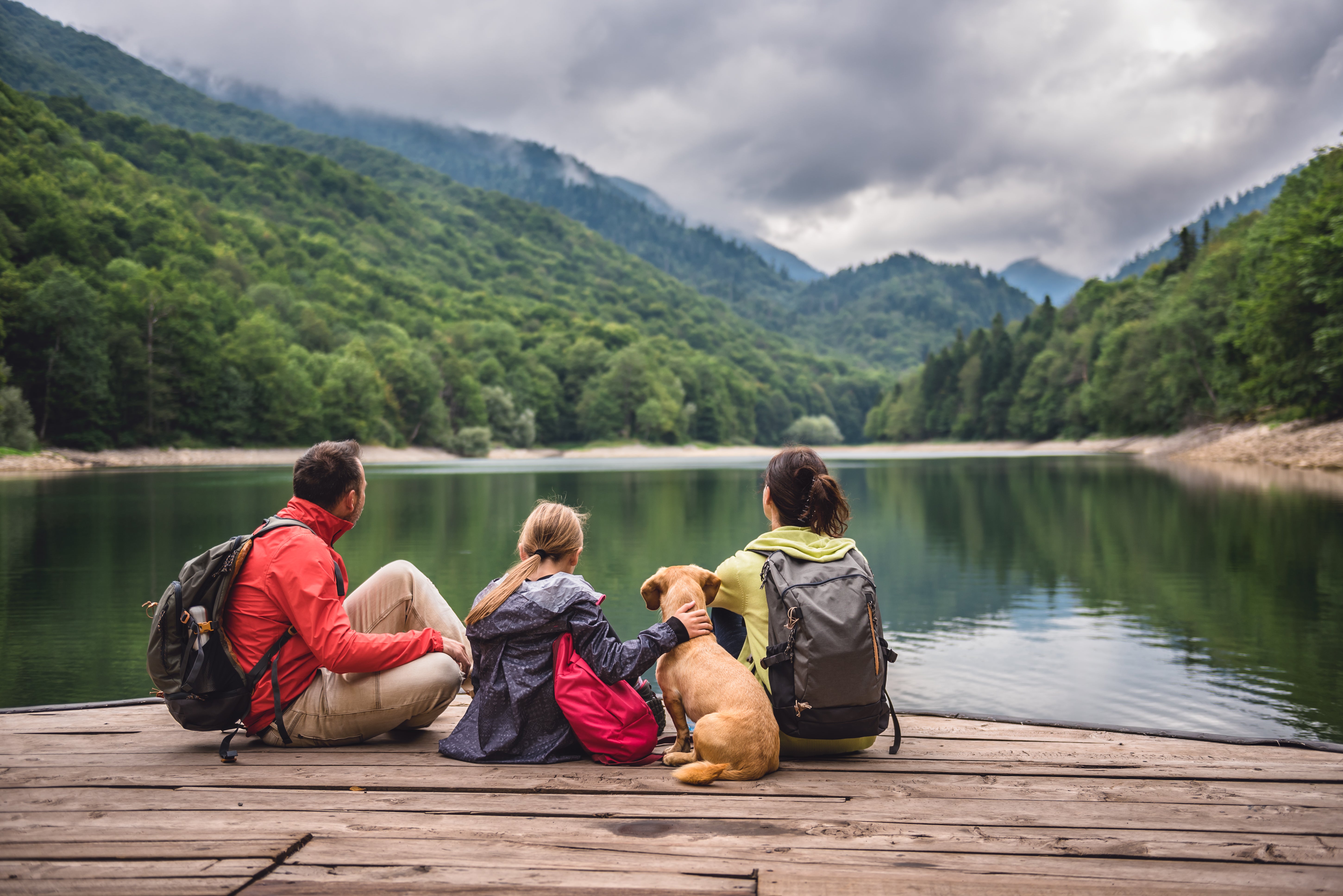 Auf dem Bild sitzt eine Familie mit einem Hund auf einem Holzsteg an einem ruhigen See, umgeben von bewaldeten Bergen. Die drei Personen – ein Mann, ein Kind und eine Frau – tragen Outdoor-Kleidung und Rucksäcke. Sie genießen gemeinsam die Aussicht auf die Natur, das spiegelnde Wasser und die grünen Hügel und Berge im Hintergrund. Die Szene wirkt entspannt und vermittelt den Eindruck eines Ausflugs oder Wanderurlaubs in der Natur.