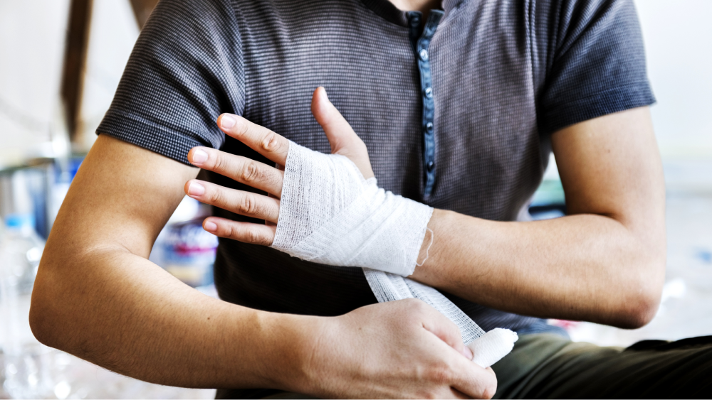 Zu sehen ist eine Person mit einem bandagierten Unterarm und einer bandagierten Hand. Die Person hält den Arm vorsichtig und ist dabei, die Bandage entweder anzulegen oder zu fixieren.