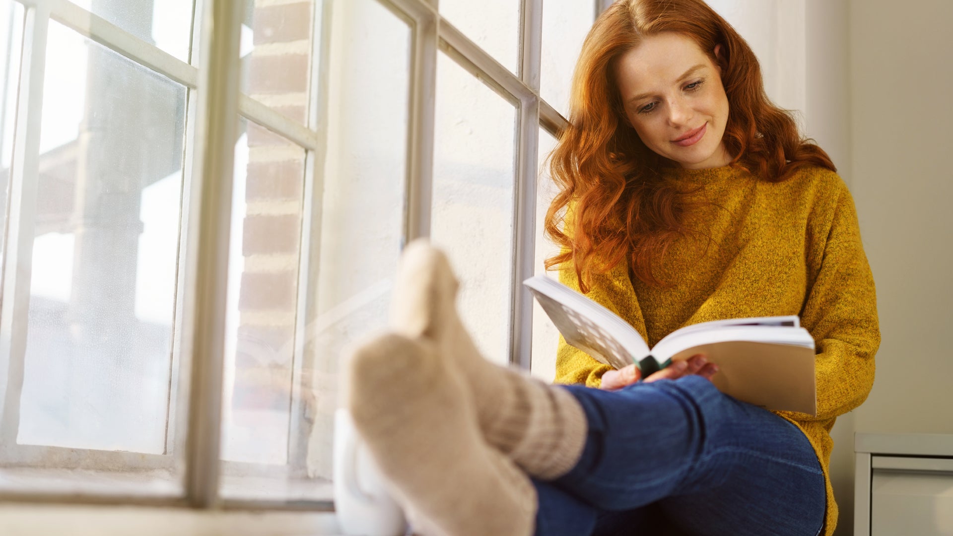 Auf dem Bild sitzt eine junge Frau entspannt an einem Fenster und liest ein Buch. Sie trägt einen gelben Pullover, Jeans und gemütliche Socken. Die Atmosphäre wirkt ruhig und behaglich, das Licht fällt sanft durch das Fenster. Es entsteht der Eindruck eines entspannten Moments, vielleicht an einem freien Tag zu Hause, an dem die Frau die Zeit zum Lesen genießt.