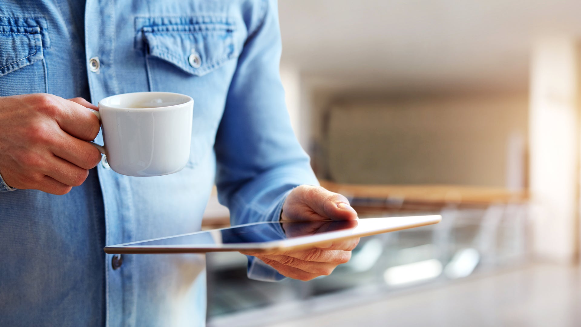 Auf dem Bild ist eine Person zu sehen, die eine weiße Kaffeetasse in der einen Hand und ein Tablet in der anderen Hand hält. Die Person trägt ein hellblaues Jeanshemd, der Kopf ist nicht im Bild. Die Szene vermittelt den Eindruck eines entspannten oder produktiven Moments, etwa beim Lesen, Arbeiten oder Surfen mit einer Tasse Kaffee, möglicherweise während einer Pause oder beim Start in den Tag.