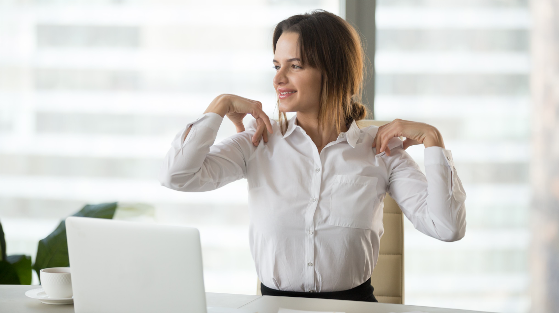 Auf dem Bild sitzt eine Frau in Business-Kleidung an einem Schreibtisch vor einem Laptop. Sie lächelt und macht eine Dehnübung, indem sie die Schultern nach hinten zieht. Die Szene spielt vermutlich in einem modernen Büro mit großen Fenstern. Die Frau wirkt entspannt und nutzt offenbar einen kurzen Moment für Bewegung während der Arbeitspause.