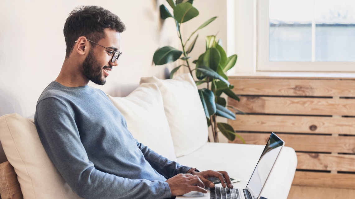Auf dem Bild sitzt ein junger Mann entspannt auf einem Sofa und arbeitet oder surft mit einem Laptop auf dem Schoß. Er trägt eine Brille und lächelt leicht, offenbar ist er konzentriert oder zufrieden bei der Arbeit. Im Hintergrund stehen eine Zimmerpflanze und ein großes Fenster, das für viel Tageslicht sorgt. Die Szene vermittelt eine angenehme, arbeits- oder freizeitorientierte Atmosphäre in einem gemütlichen Zuhause.