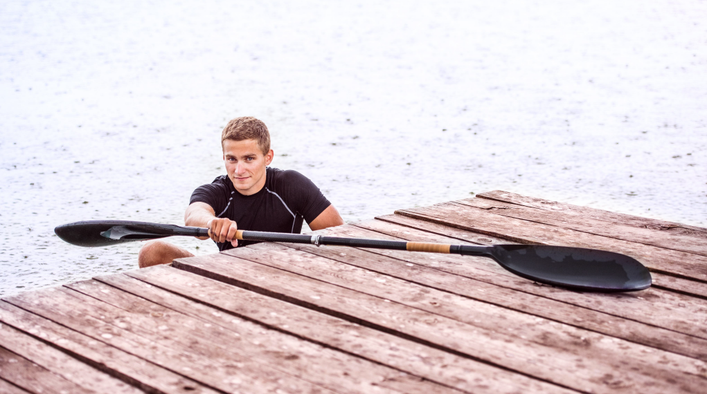 Auf dem Bild ist ein junger Mann zu sehen, der im Wasser neben einem hölzernen Steg schwimmt oder sich daran festhält. Er hat ein Paddel in der Hand, das über den Steg gelegt ist. Der Hintergrund zeigt eine große Wasserfläche. Die Szene wirkt sportlich und es sieht so aus, als hätte er zuvor gepaddelt oder gekajakt und ist nun kurz am Steg angehalten oder ins Wasser gestiegen.