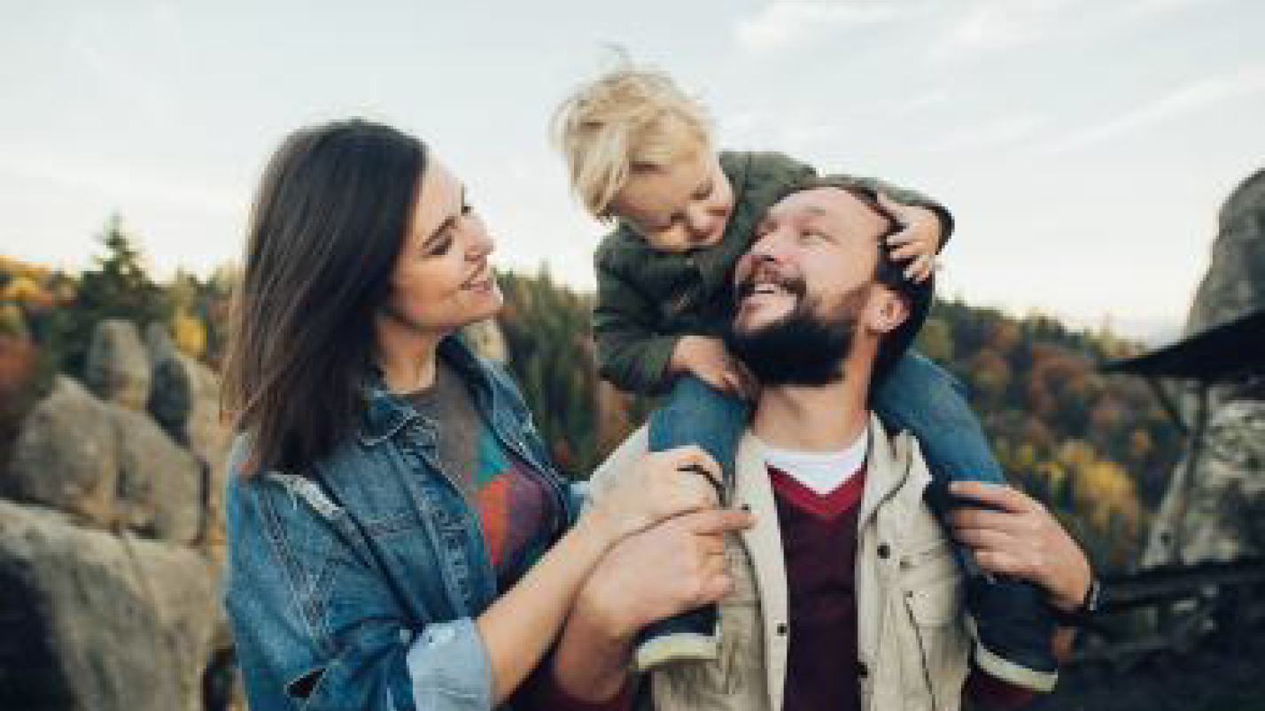 Auf dem Bild ist eine fröhliche Familie in der Natur zu sehen. Ein Mann trägt ein Kind auf den Schultern, während die Frau neben den beiden steht. Alle drei lächeln sich glücklich an und wirken sehr verbunden. Im Hintergrund erkennt man Felsen und Wälder, was auf einen Ausflug oder eine Wanderung in einer landschaftlich schönen Gegend hindeutet. Die Szene strahlt Freude, Zusammenhalt und Familienglück aus.