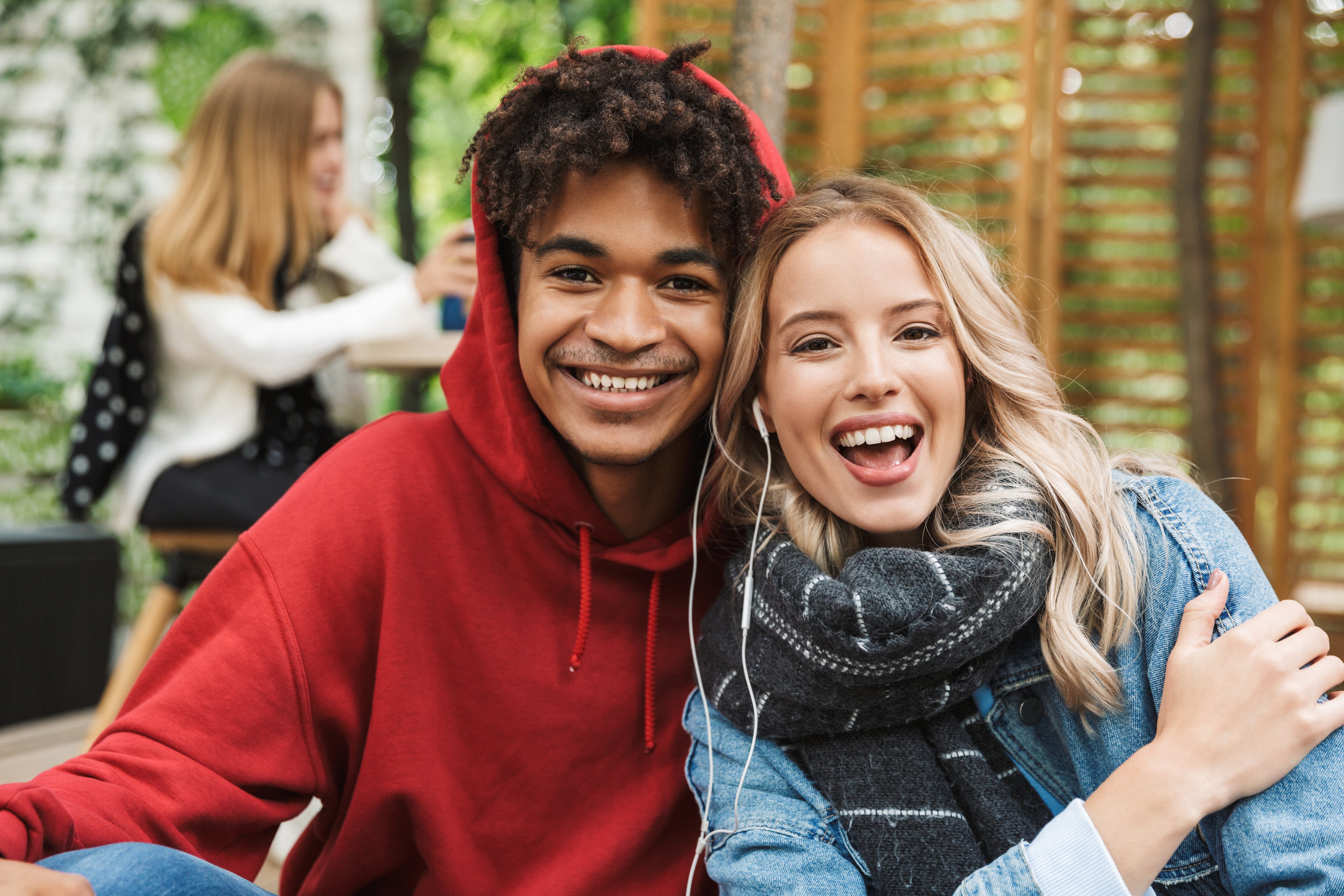 Auf dem Bild sind zwei junge Menschen zu sehen, die eng nebeneinander sitzen, lächeln und direkt in die Kamera schauen. Der junge Mann trägt einen roten Hoodie, die junge Frau einen Schal. Sie wirken fröhlich und entspannt. Beide teilen sich ein Paar Ohrhörer, was auf gemeinsame Musik oder ein Video hindeutet. Im Hintergrund sitzt eine weitere Person verschwommen an einem Tisch. Die Szene vermittelt Freundschaft, Freude und ein gemeinsames, entspanntes Beisammensein.