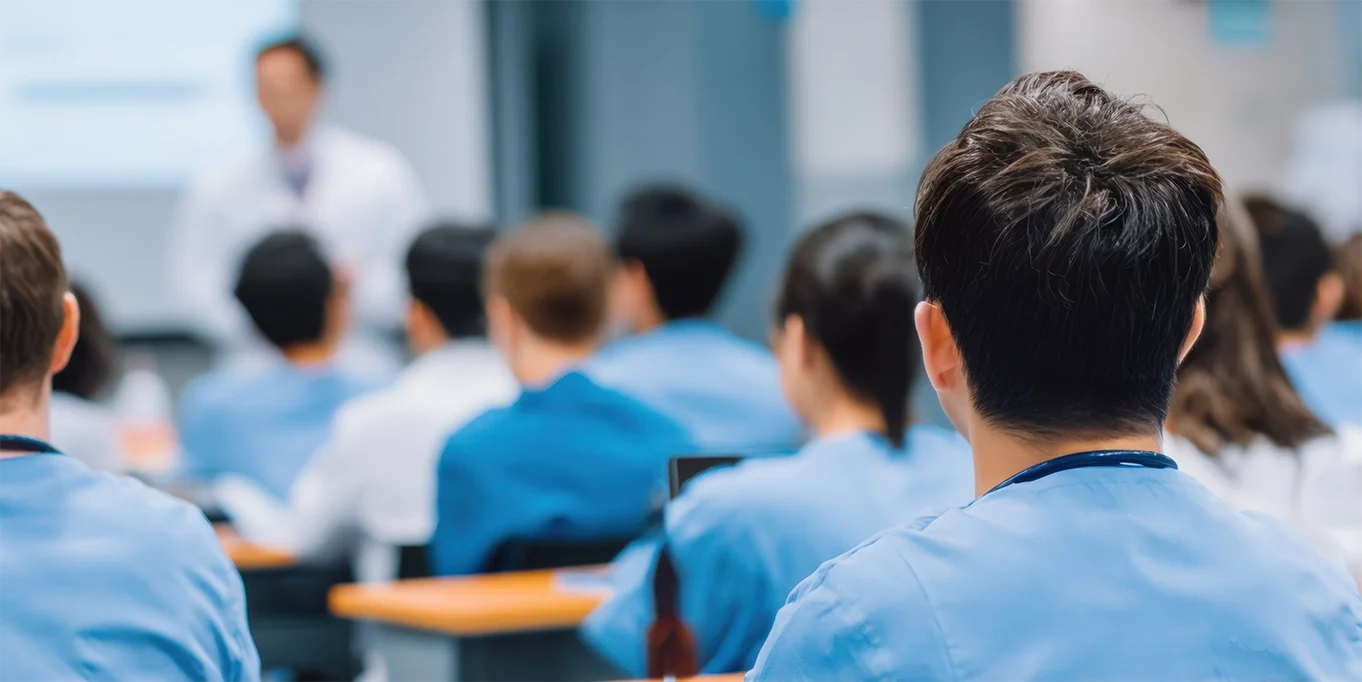 Mehrere Personen in hellblauer medizinischer Arbeitskleidung sitzen in einem Seminar- oder Schulungsraum an Tischen und sind mit dem Rücken zur Kamera ausgerichtet.