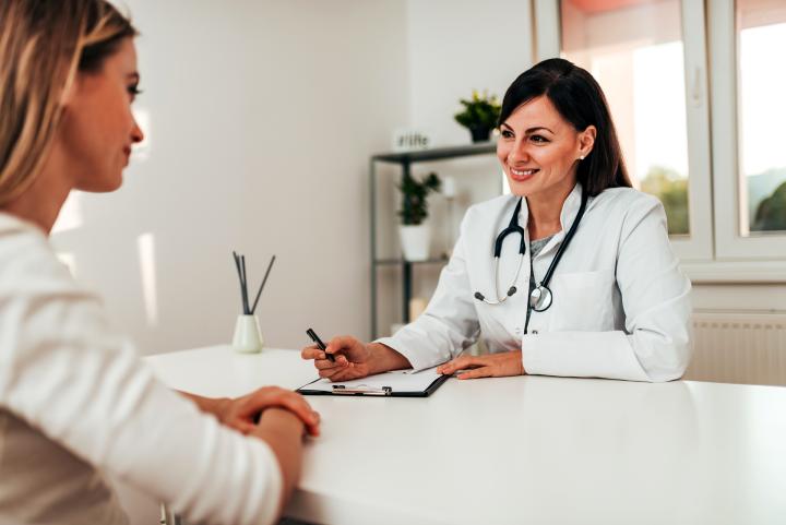 Eine Ärztin sitzt an einem Schreibtisch und führt ein Gespräch mit einer Patientin. Die Ärztin trägt einen weißen Kittel und hat ein Stethoskop um den Hals, sie lächelt und hält einen Stift sowie ein Klemmbrett in der Hand. 