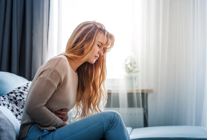 Eine junge Frau mit langen Haaren sitzt auf einem Bett oder Sofa in einem hellen Raum. Sie hält sich mit beiden Händen den Bauch und wirkt, als hätte sie Schmerzen oder Unwohlsein.