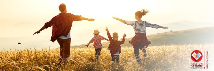 Eine Familie mit zwei Erwachsenen und zwei Kindern, laufen fröhlich durch ein sonniges Getreidefeld. Alle haben die Arme ausgebreitet und bewegen sich gemeinsam in die Weite der Natur. In der rechten unteren Ecke befindet sich ein Logo mit der Aufschrift „Passion für seltene Erkrankungen – Hämostaseologie“. Die Stimmung ist lebhaft und unbeschwert.