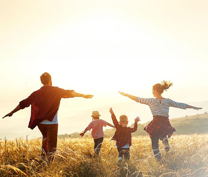 Eine Familie mit zwei Erwachsenen und zwei Kindern, laufen fröhlich durch ein sonniges Getreidefeld. Alle haben die Arme ausgebreitet und bewegen sich gemeinsam in die Weite der Natur. In der rechten unteren Ecke befindet sich ein Logo mit der Aufschrift „Passion für seltene Erkrankungen – Hämostaseologie“. Die Stimmung ist lebhaft und unbeschwert.